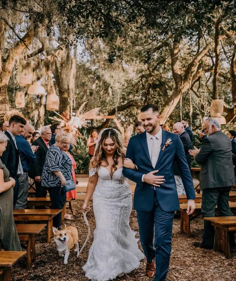 Newlyweds walk back up the aisle under oak trees as guests cheer, a corgi trotting beside the bride
