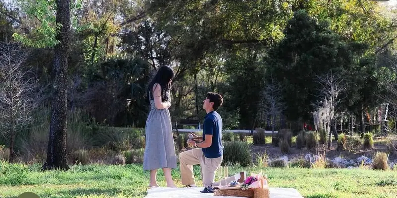 A man kneels to propose to a woman beside a picnic set up on a lawn edged with trees