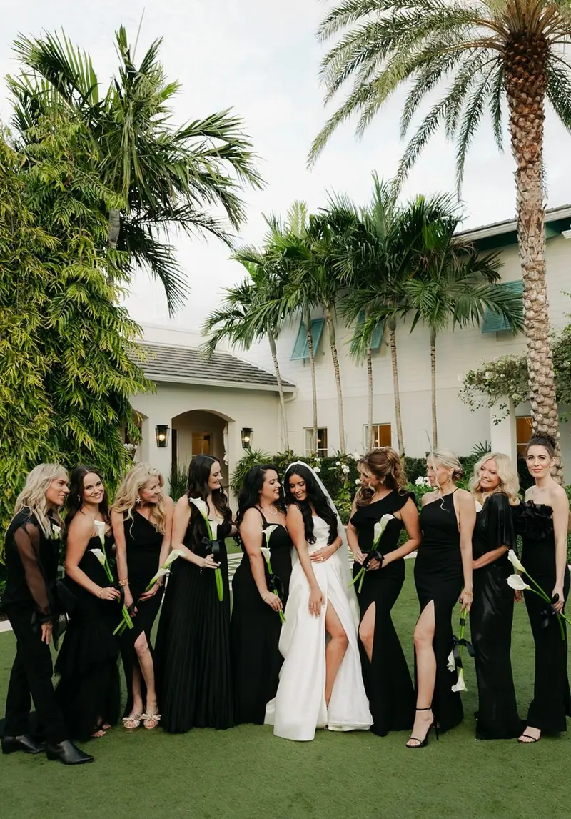A bride stands with her bridesmaids in long black dresses on a lawn lined with palm trees