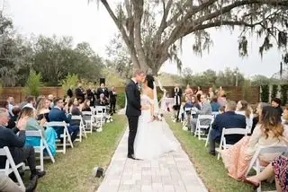 A couple kisses at the end of a brick aisle under a large oak tree, guests seated on both sides