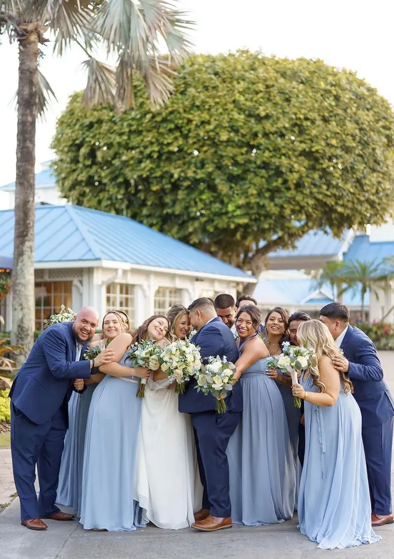 A bride and her wedding party in dusty blue dresses and navy suits squeeze together for a group hug