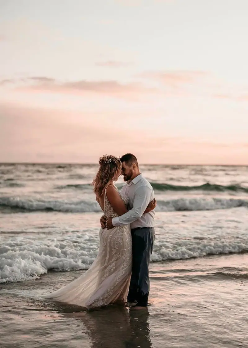 A couple embraces in the ocean surf at sunset, the bride's dress trailing in the water