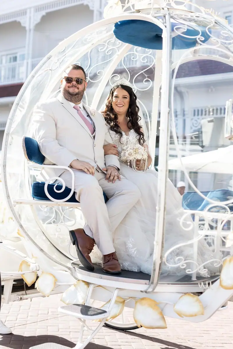 A bride and groom sit in a white Cinderella-style carriage, the groom in a cream suit and sunglasses