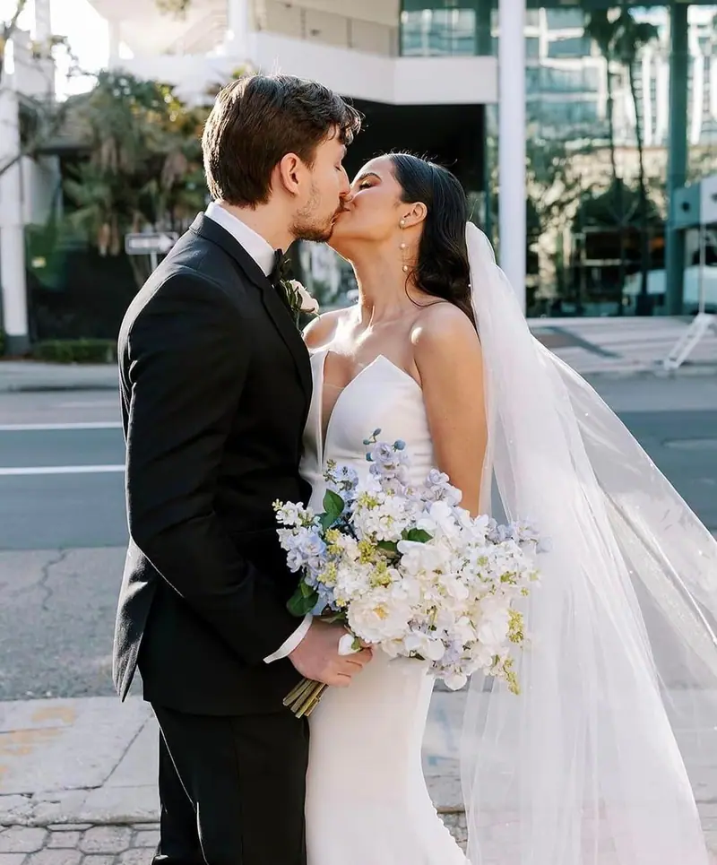 A bride and groom kiss on a city sidewalk, the bride holding a bouquet of white and blue flowers