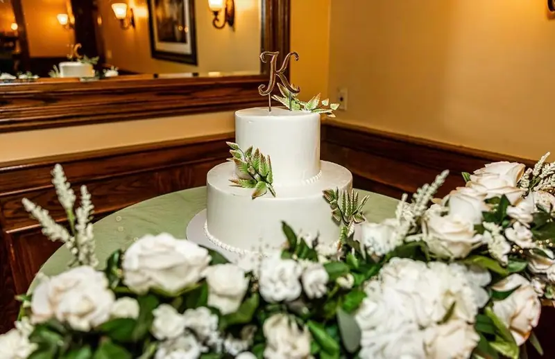 A two-tier white wedding cake with greenery and a monogram topper, ringed by white roses