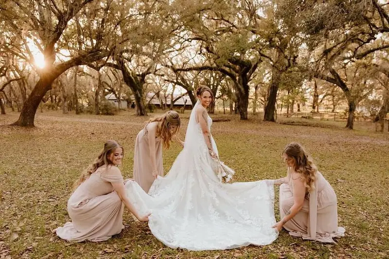 Two bridesmaids in blush dresses spread the bride's lace train under oak trees at golden hour