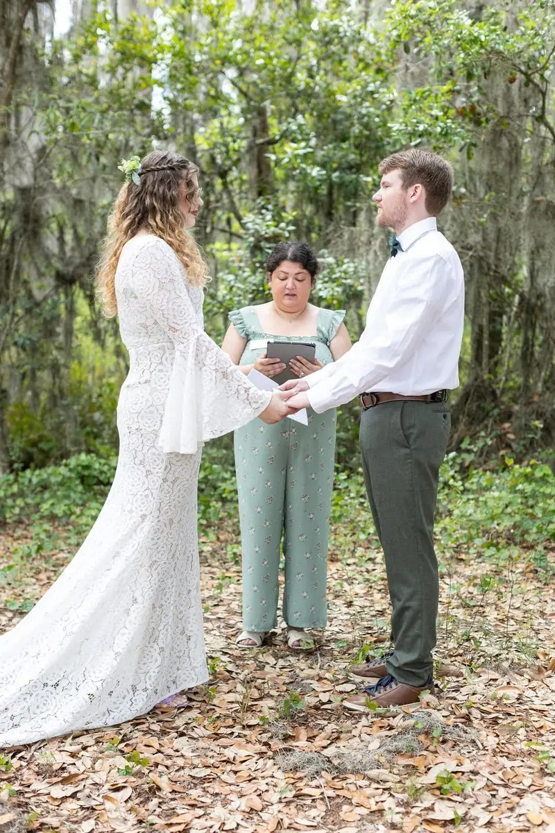 A couple holds hands as an officiant reads during a woodland ceremony under mossy trees