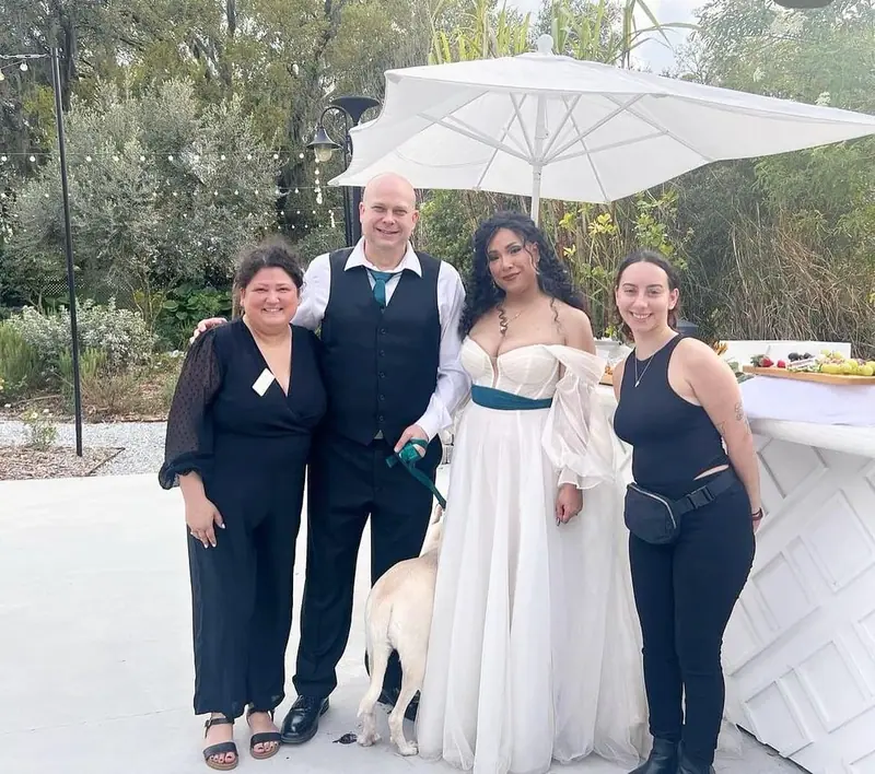 A bride and groom pose with two women in black and a yellow Labrador beside a white umbrella