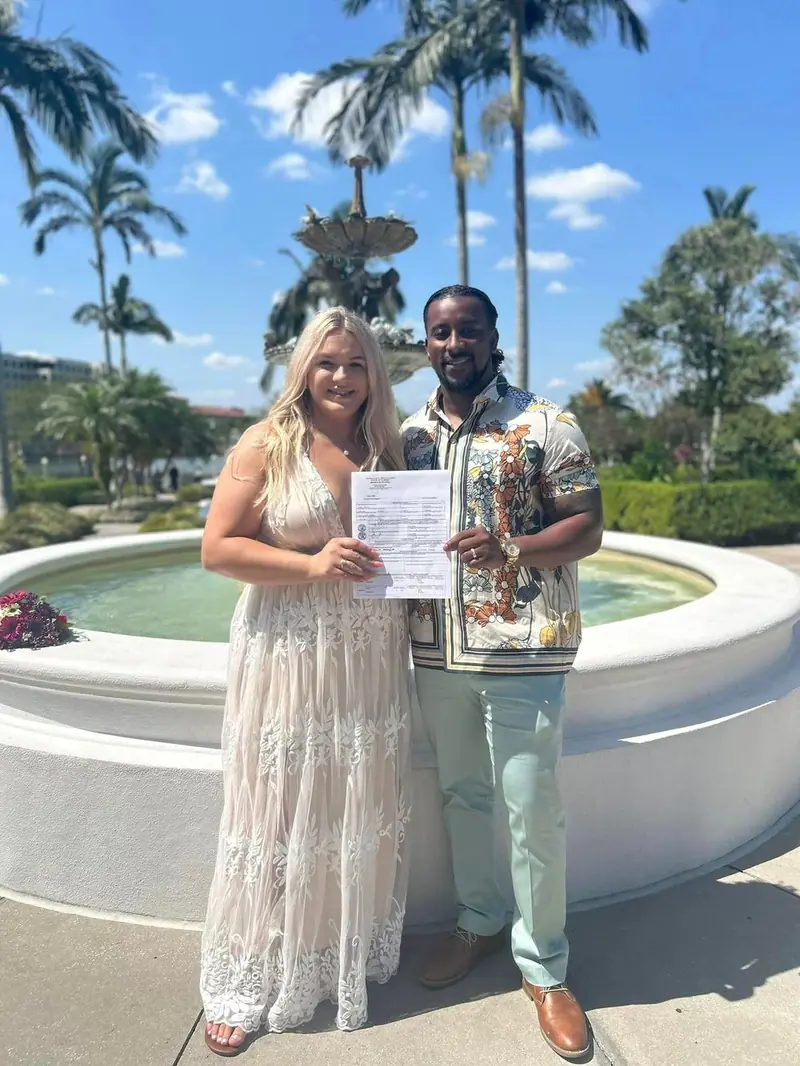 A smiling couple holds up their signed marriage license beside a fountain under palm trees