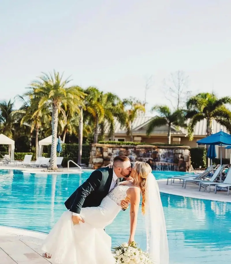A groom dips his bride for a kiss beside a resort pool lined with palm trees