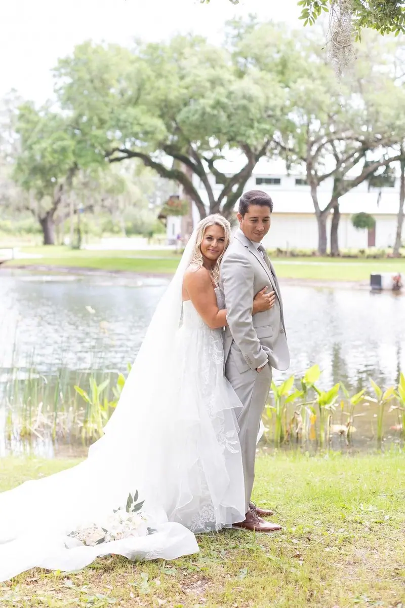 A bride and groom embrace beside a pond and oak trees, her veil spread across the grass