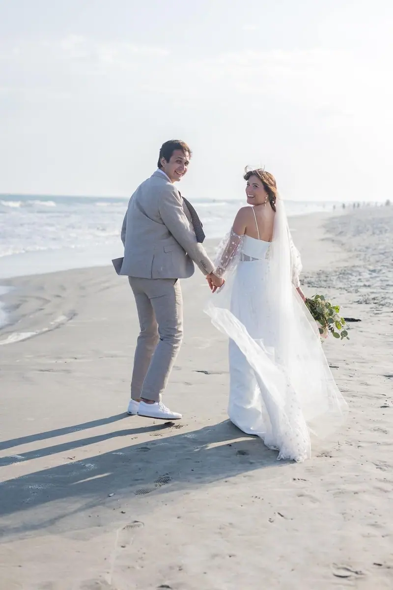 A couple walks hand in hand along the beach, looking back over their shoulders