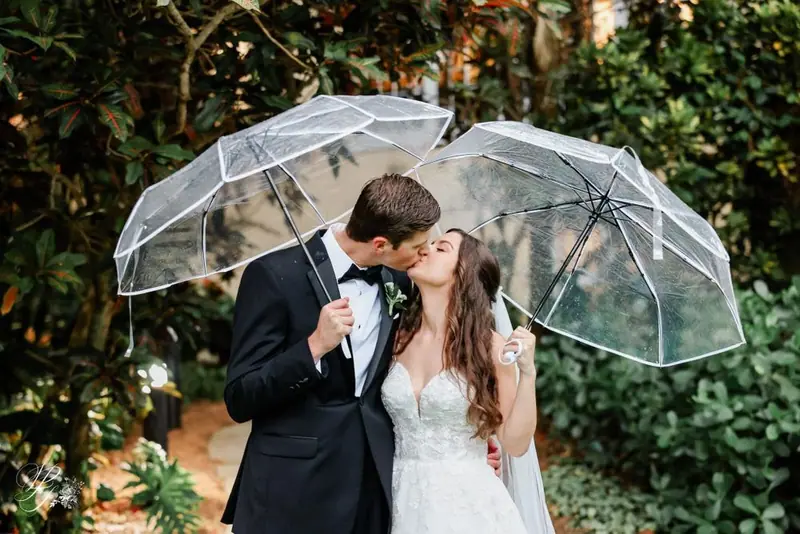 A bride and groom kiss under two clear umbrellas in a leafy garden