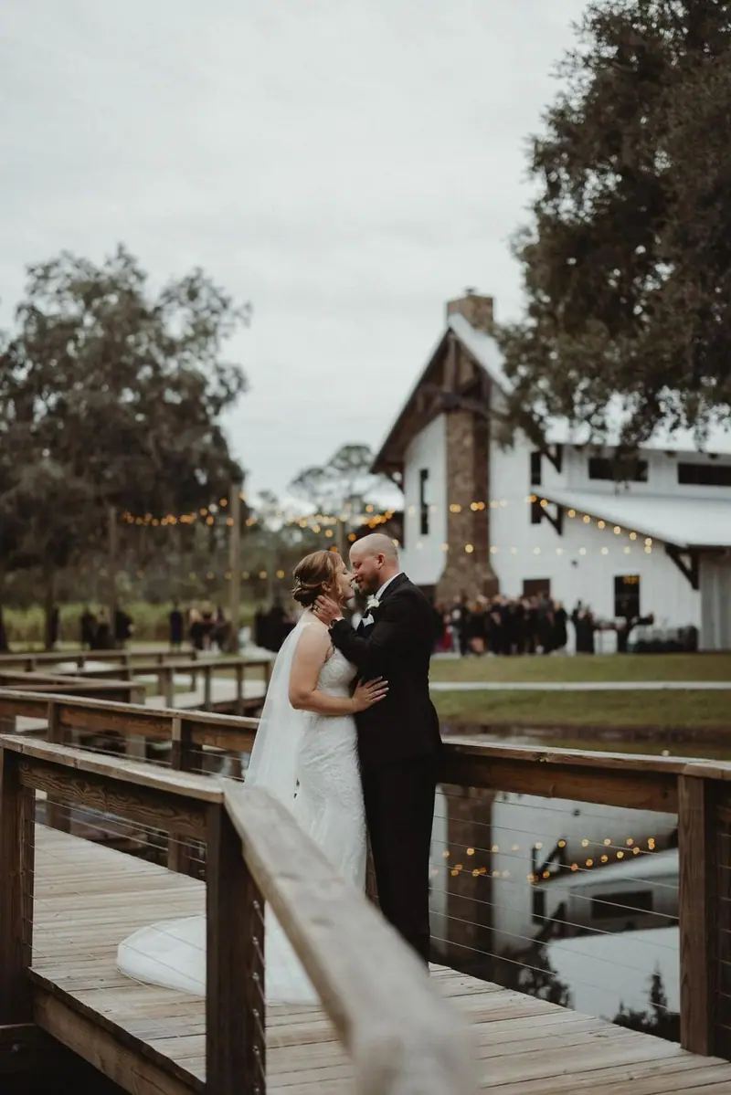 A bride and groom embrace on a wooden dock at dusk, string lights and a white barn behind them