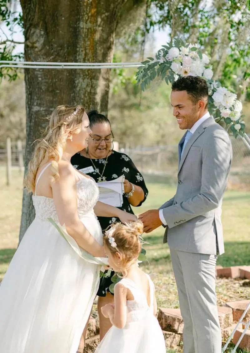 A young girl in white stands between the bride and groom as the officiant reads under a large tree