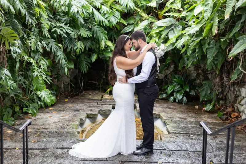 A bride and groom embrace in a stone courtyard framed by large tropical leaves
