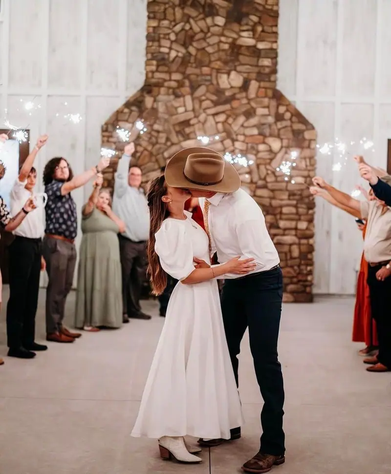 A couple in a white dress and a cowboy hat kiss during a sparkler send-off in a white barn venue