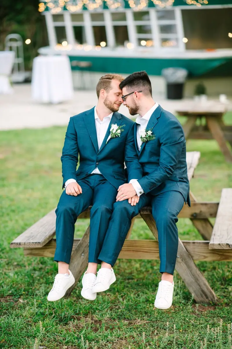 Two grooms in teal suits sit on a picnic table with their foreheads touching