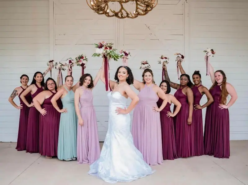 A bride raises her bouquet with her bridesmaids in plum, lavender and sage dresses
