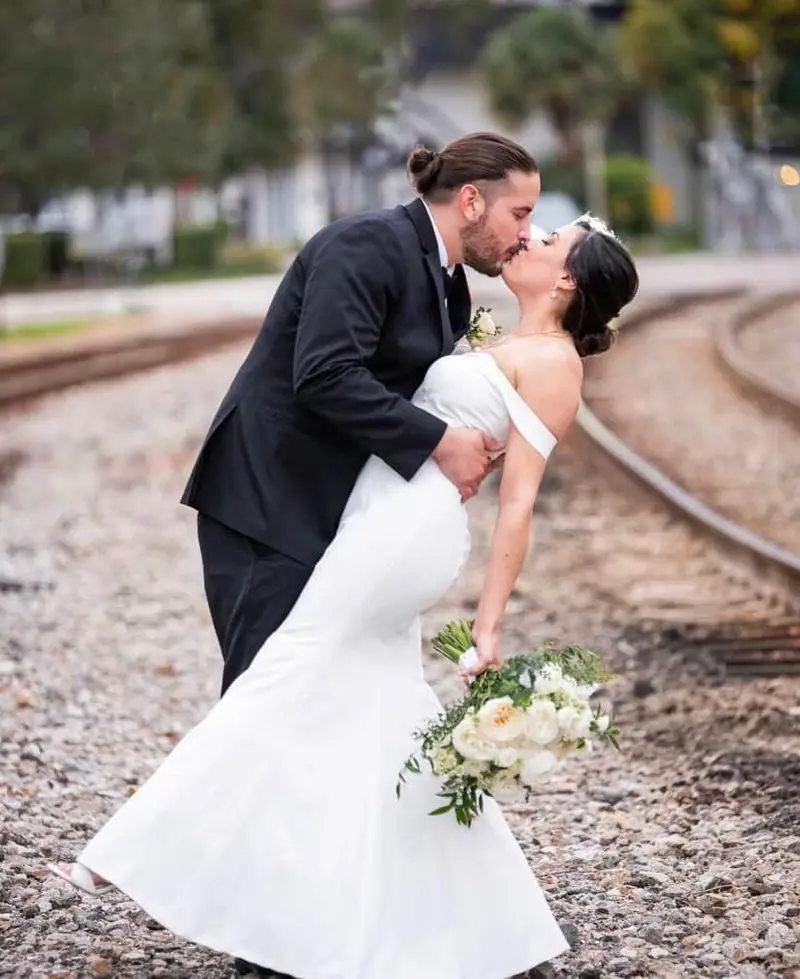 A groom dips his bride for a kiss on railroad tracks, her bouquet of white flowers in hand