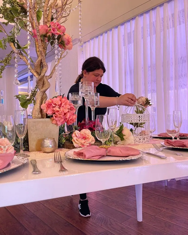 A woman arranges pink flowers and place settings on a reception table under purple lighting