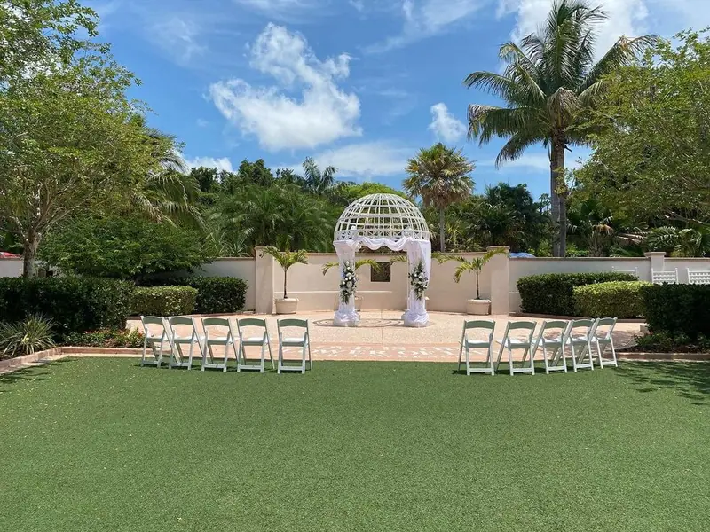 White chairs set on a lawn facing a white gazebo under palm trees and blue sky