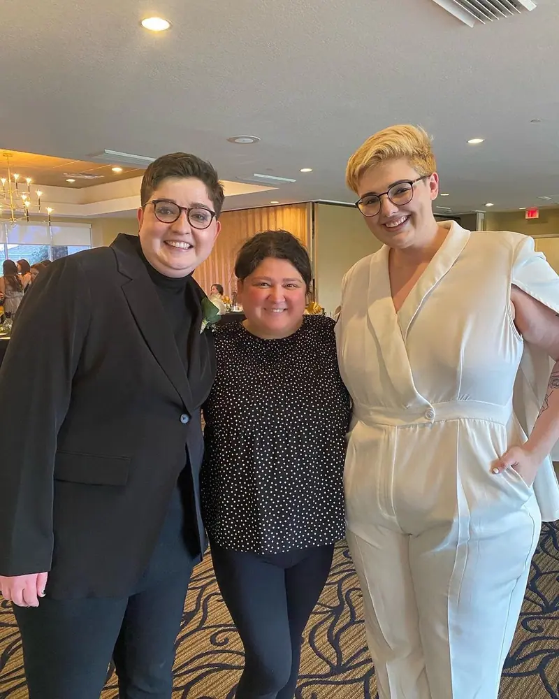 A couple, one in a black suit and one in a white jumpsuit, smiles with a woman between them