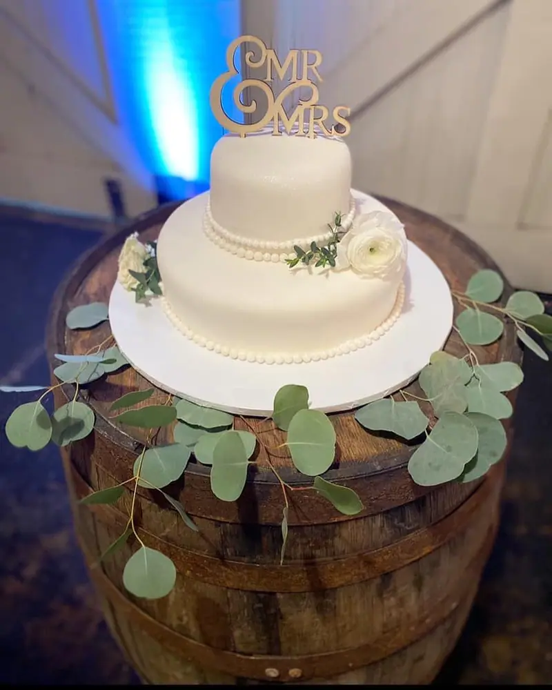 A two-tier white wedding cake with a Mr & Mrs topper on a wooden barrel wrapped in eucalyptus