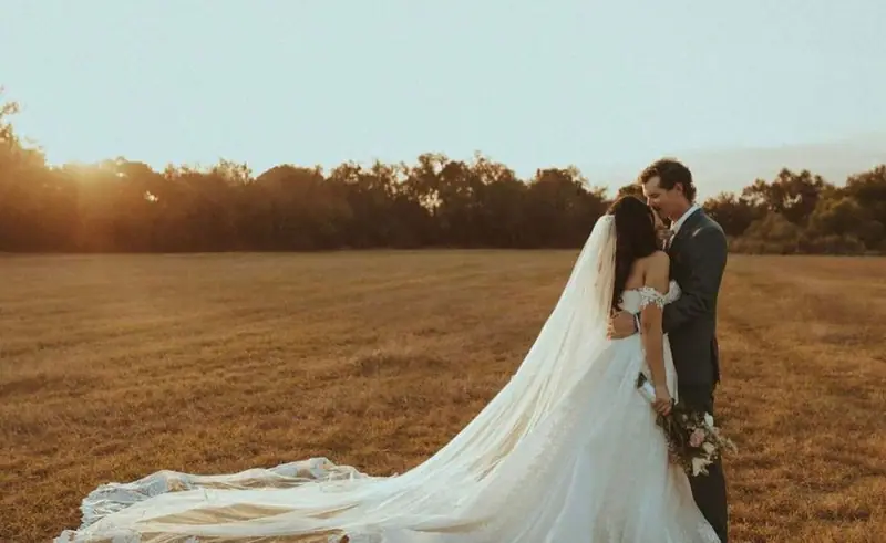 A couple kisses in a golden field at sunset, the bride's long veil trailing across the grass