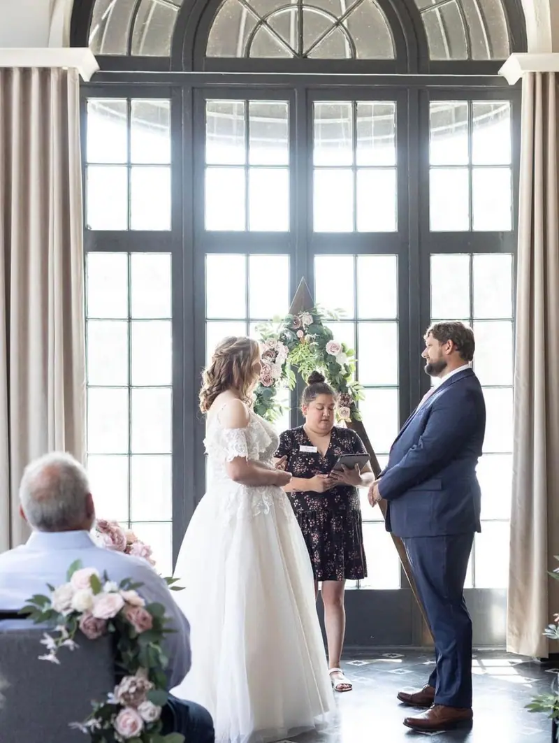 A couple faces each other before an officiant in front of tall arched windows
