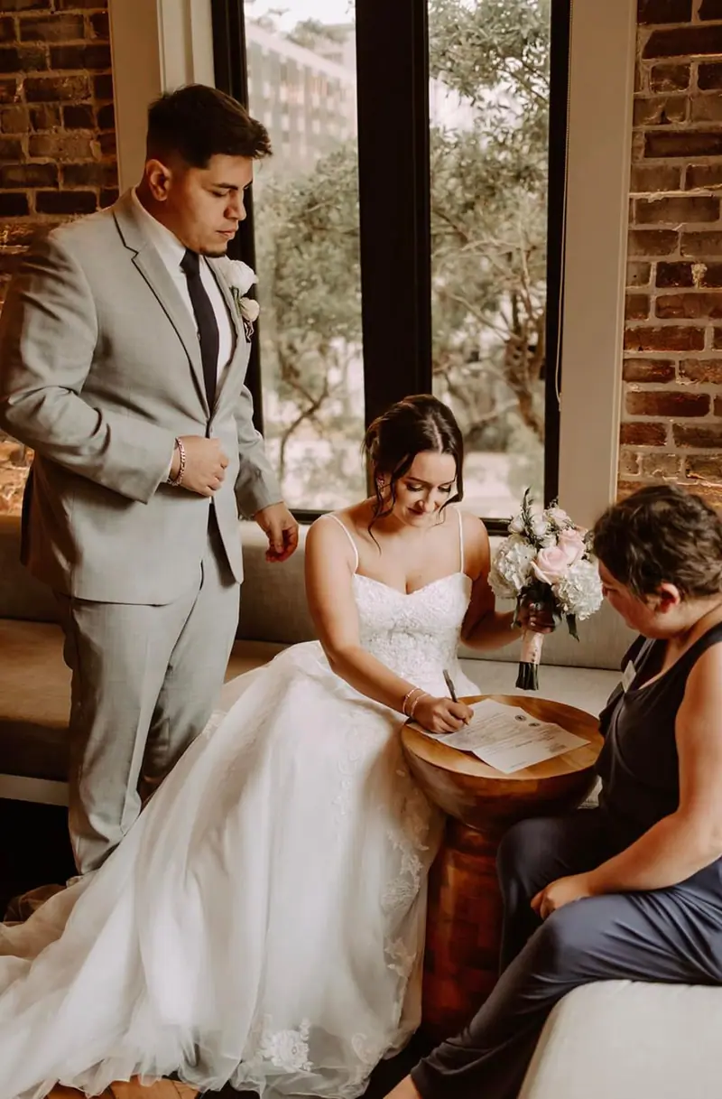 A bride signs the marriage license as the groom and a woman look on