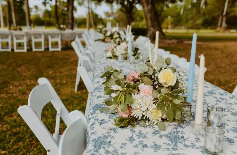 A long outdoor table with a blue and white floral tablecloth, flowers, candles and white chairs