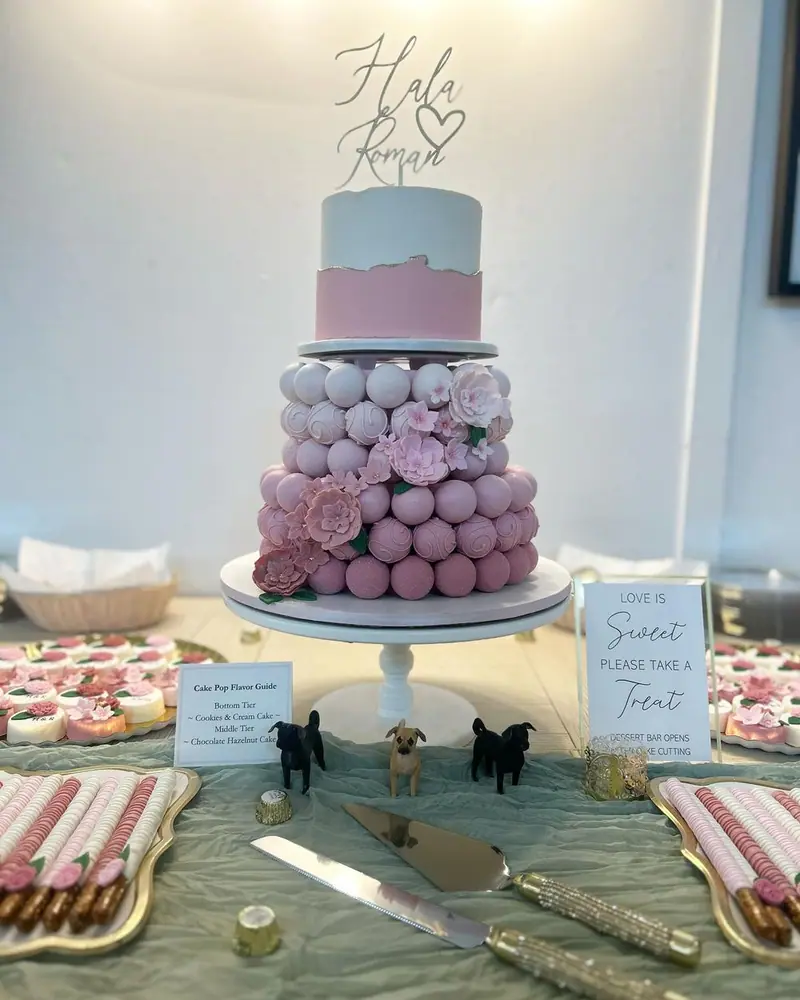 A pink and white tiered cake with a tier of cake pops, on a dessert table with treats
