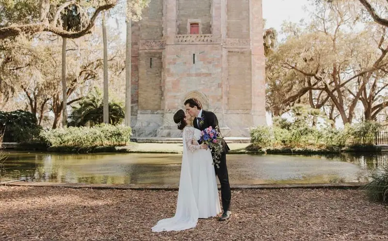 A couple kisses by a reflecting pool in front of a historic stone tower framed by oak trees