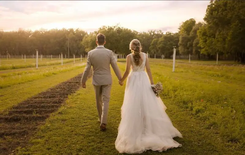 A bride and groom walk hand in hand across a field toward the trees at sunset