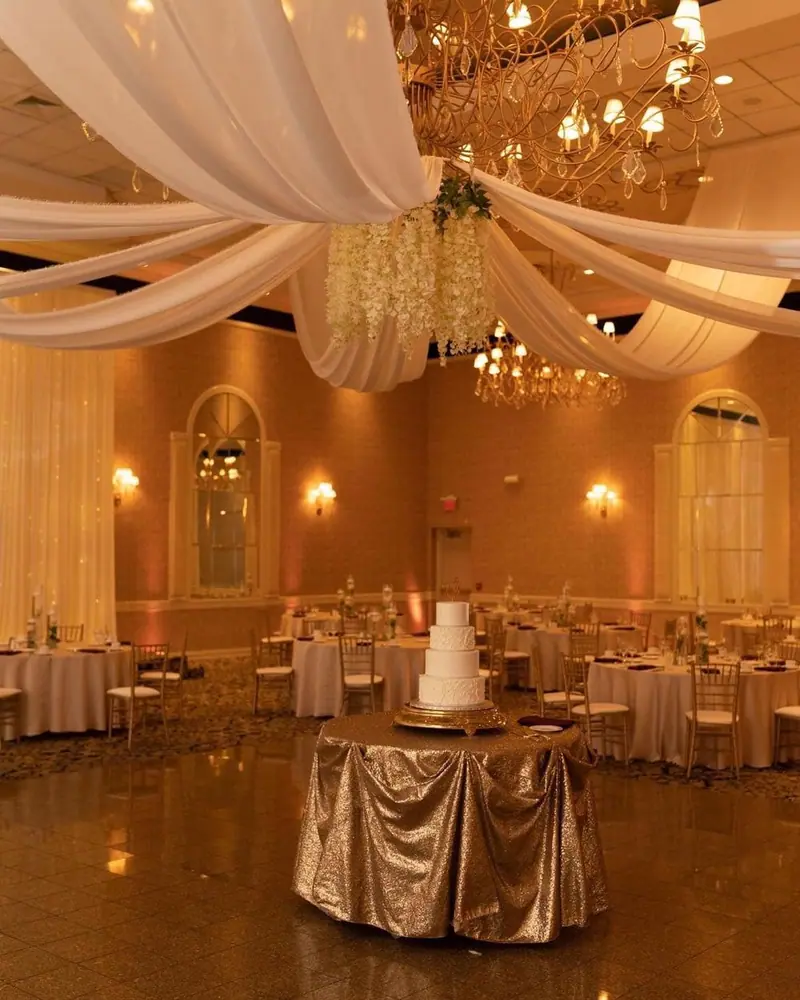 A ballroom with sheer white ceiling drapes, chandeliers and gold tablecloths, the wedding cake in the center