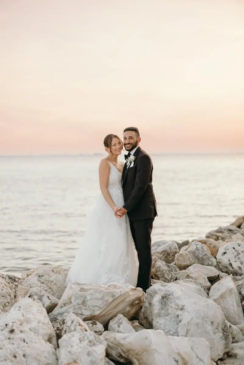 A bride and groom embrace on rocks at the water's edge at sunset